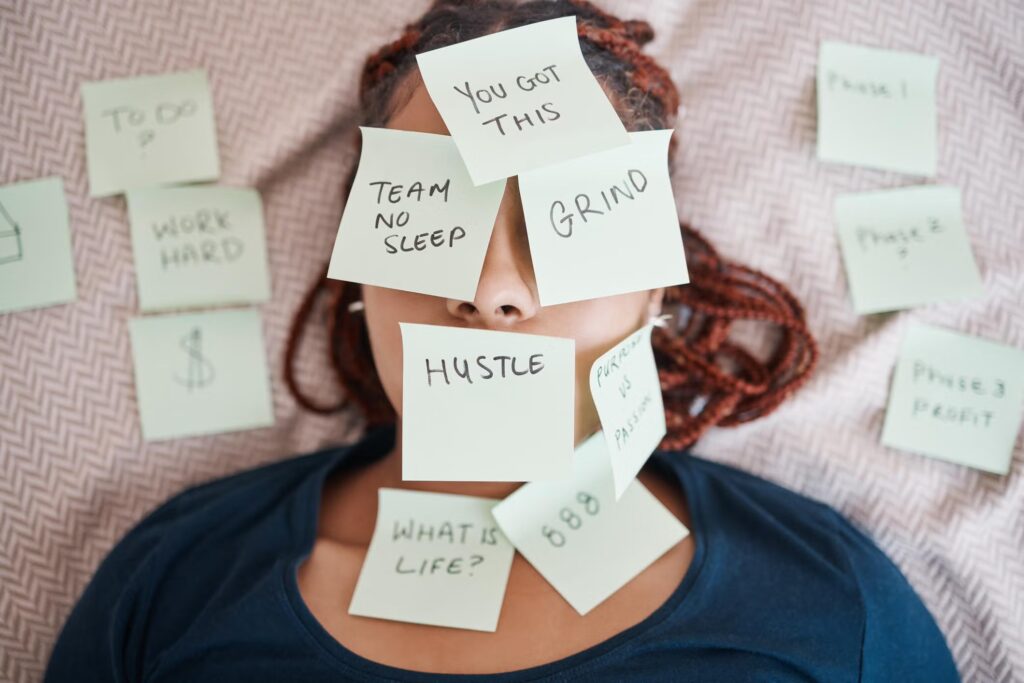 A woman lies on a bed covered in sticky notes that read: "You got this," "Grind," "Hustle," and "Team No Sleep." 
